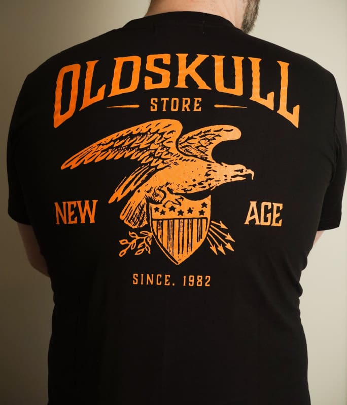 This shirt is similar to the Harley Davidson style of shirts.  It is black with orange print.  It has a small Oldskull Shirts logo on the front.  On the back it has an large eagle over a shield design with the words Oldskull Store written over it.  It is a vintage streetwear design. 